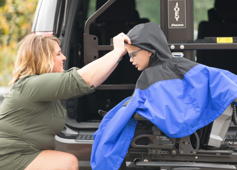 Mother helping a child put on a wheelchair poncho in front of an open vehicle